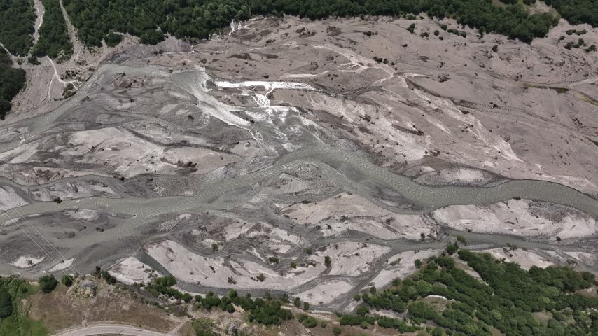 Aerial view of the wide, sediment-filled riverbed with winding channels and surrounding green forests near Chiora, Racha-Lechkhumi and Lower Svaneti, Georgia.