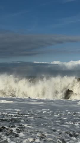 Vertical video of powerful sea waves crashing on a pebble beach with white foam under a blue sky. Ocean coast nature footage.⁠
