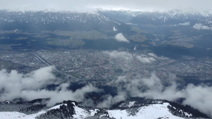 High aerial view of Innsbruck spread across the Inn Valley during light snowfall, framed by snowy tree covered slopes below and an alpine mountain range in the background, Nordkette, Tyrol, Austria