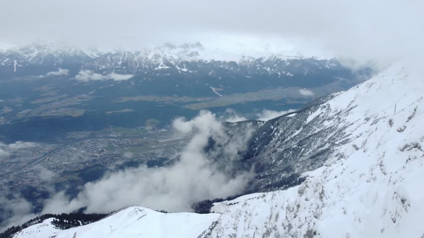Aerial view over a snowy Nordkette ridge as low clouds pour down a forested gully toward Innsbruck and the Inn Valley, with snowcapped Alps beyond, Tyrol, Austria