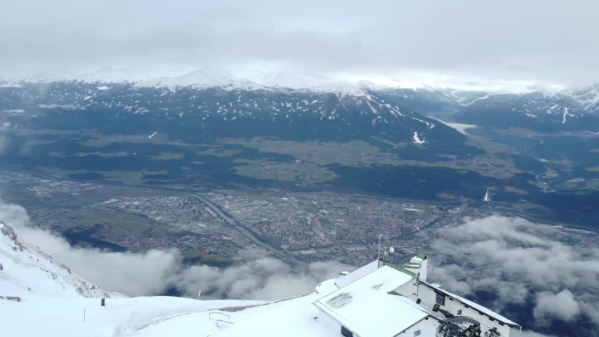 High aerial panoramic view of Innsbruck from the top of the snow-covered Hafelekar cable car station high on the Nordkette, looking down past drifting clouds to the city of Innsbruck spread across the Inn Valley, with a flock of birds flying just below, Tyrol, Austria