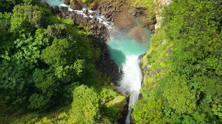 High angle top view as camera slowly moves forward over massive Victoria Falls plunging into turquoise pool surrounded by lush green tropical forest in Dominica