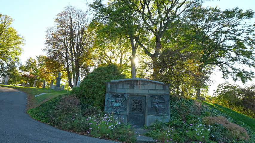 A mausoleum sits in a planted slope beside a curving road. Sunlight filters through trees above relief panels and flowers surrounding a gray entrance.