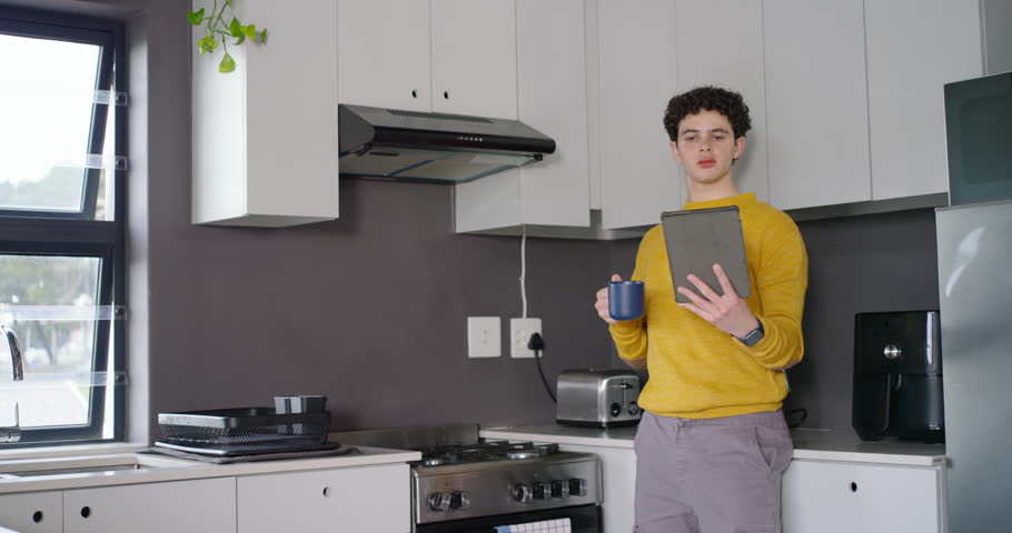 African American man leaning in kitchen with tablet and mug, processing tablet content and reacting. Device, beverage, home, window, plant, daylight, modern