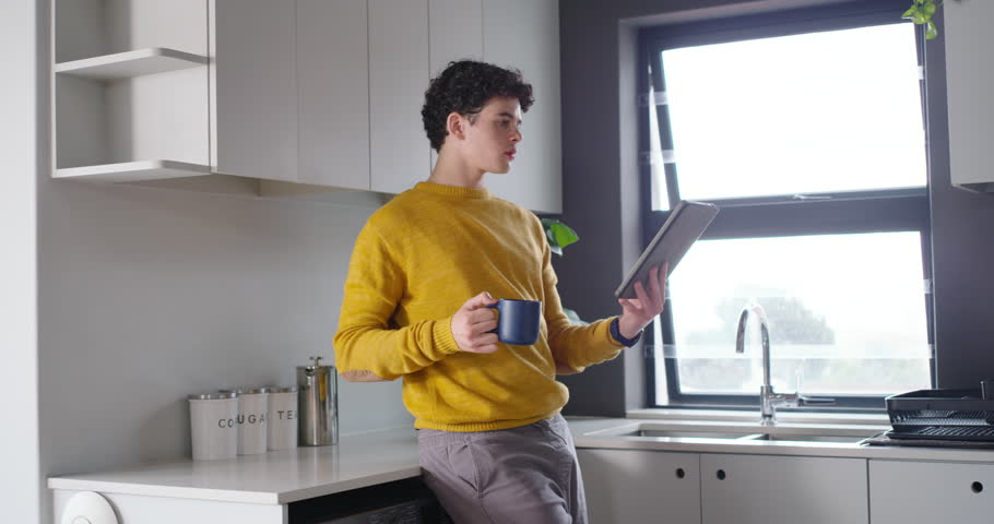 African man 20s leaning on kitchen counter reading tablet, smiling, sipping mug; sun prompts glance. Home, lifestyle, modern, daylight, coffee, device, candid