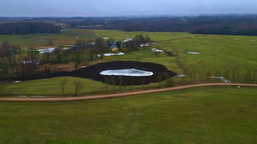 Aerial view of a small frozen pond surrounded by dark soil in a green field with a dirt road and distant trees in Latvia.
