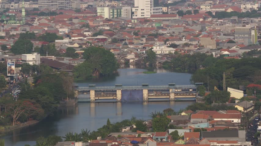 High-angle view of a river dam and sluice gate in Surabaya surrounded by residential houses and dense urban buildings in East Java, Indonesia.