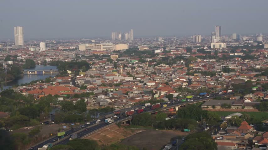 Aerial view of Surabaya city skyline with highway traffic, bridge over river, and densely packed residential roofs during golden hour sunset in East Java, Indonesia.