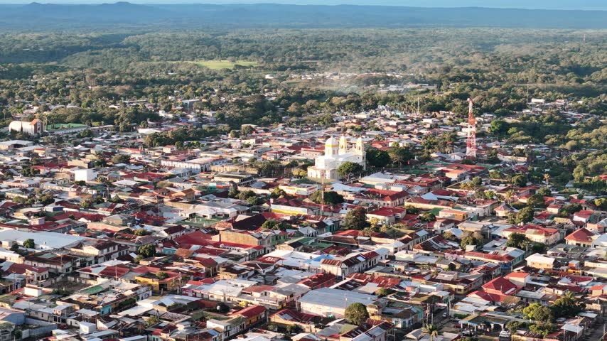 An aerial view looks over a town with homes, shops, roads, and hills in the distance during daylight.