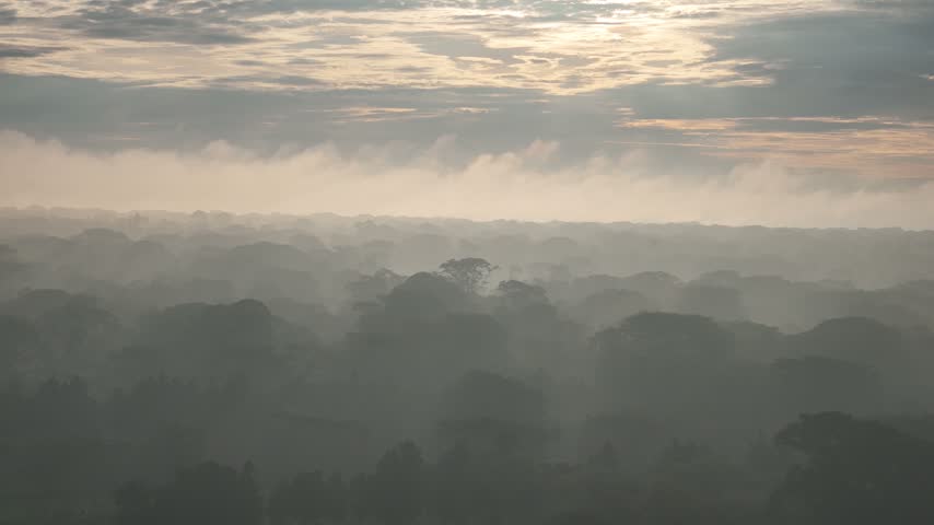 Morning light reaches across forested mountain ridges beneath a cloudy sky, with mist hanging over the hills.