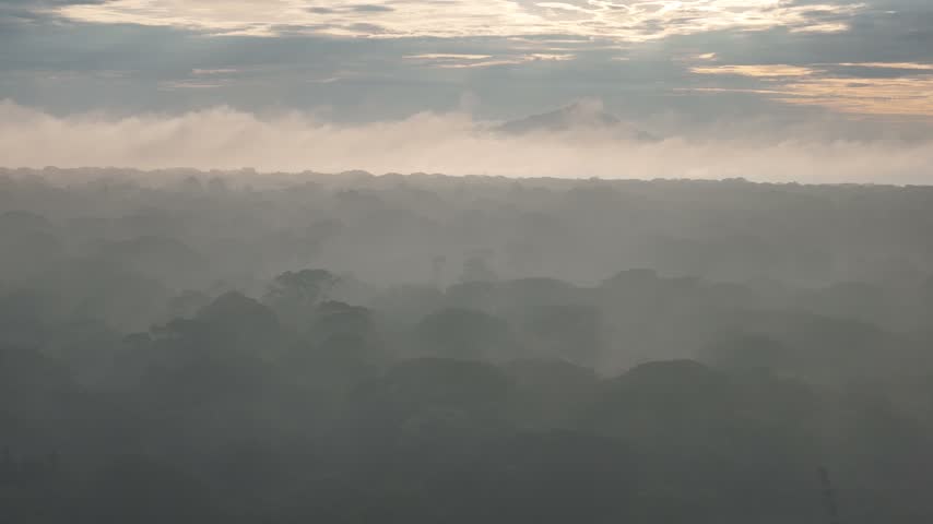 Mist covers forested mountain ridges at sunrise, with layers of hills visible beneath a cloudy sky.