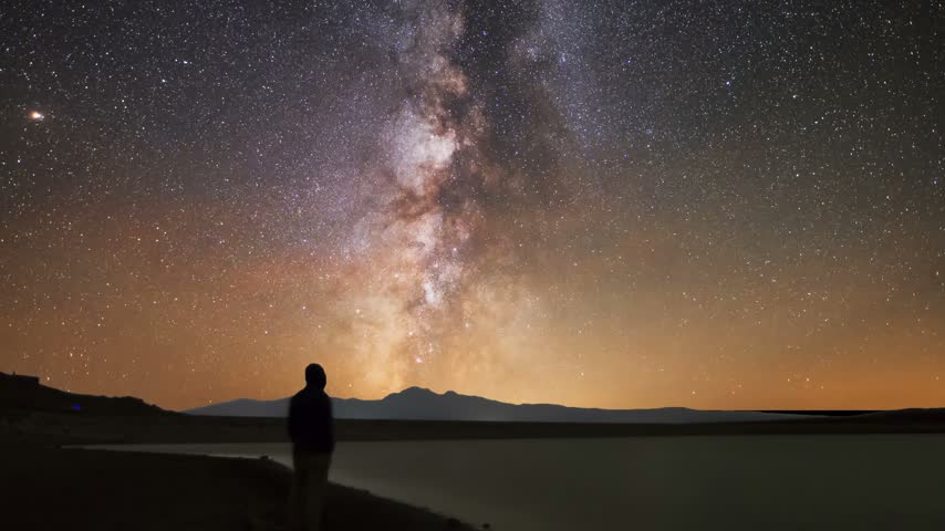 A human silhouette stands on the shore, gazing at the mountains and the lake beneath a brilliant Milky Way. A starry sky.