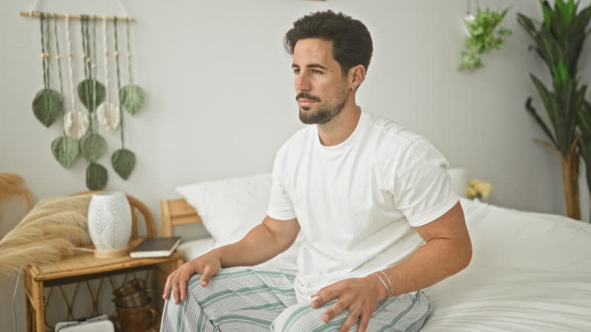 Man grimacing and clutching lower back while sitting on bed inside a residential building with clear backache; pain recovery.
