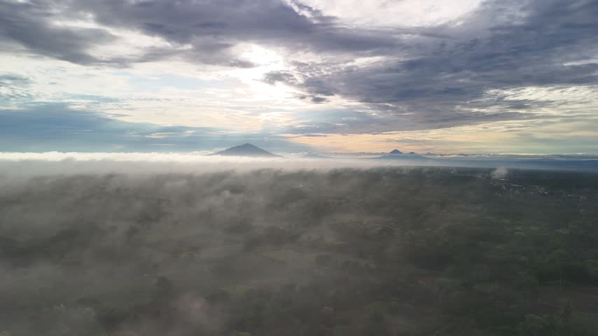 Clouds move over a forest near distant mountain peaks beneath a cloudy sky during the day.