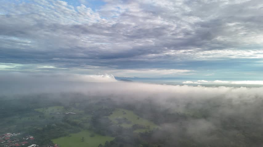 Low clouds cover fields and homes across a rural landscape beneath a cloudy sky during the day.