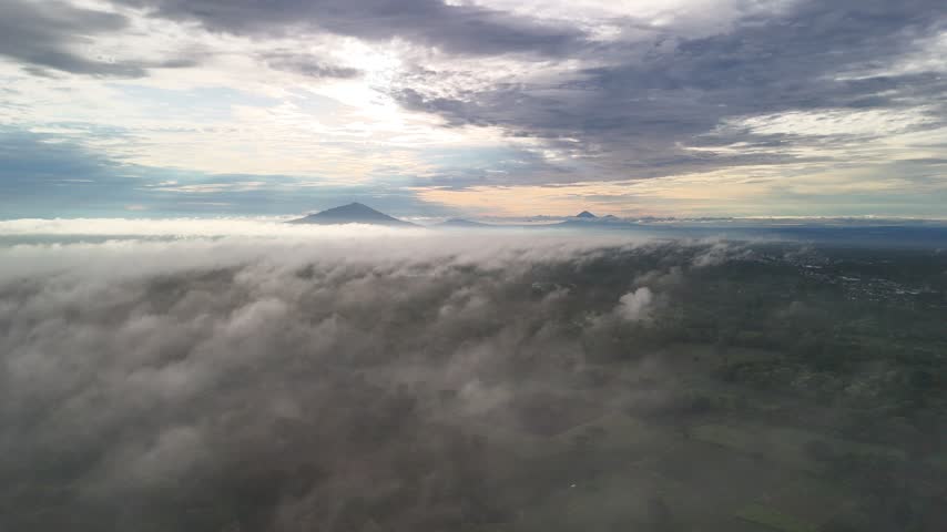 Clouds cover the land around mountain peaks at sunrise, viewed from above in the early morning.