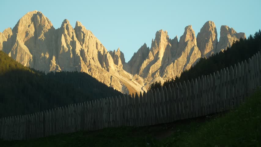 Dolomites jagged limestone peaks bathed in warm late afternoon sun above dark forest with weathered wooden palisade fence in foreground, panoramic alpine landscape scene