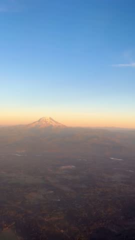 Aerial view of snow-capped Mount Rainier in Washington State at sunset, with warm golden light illuminating the mountain and surrounding landscape beneath a clear blue sky. Vertical 4K footage filmed from an airplane.