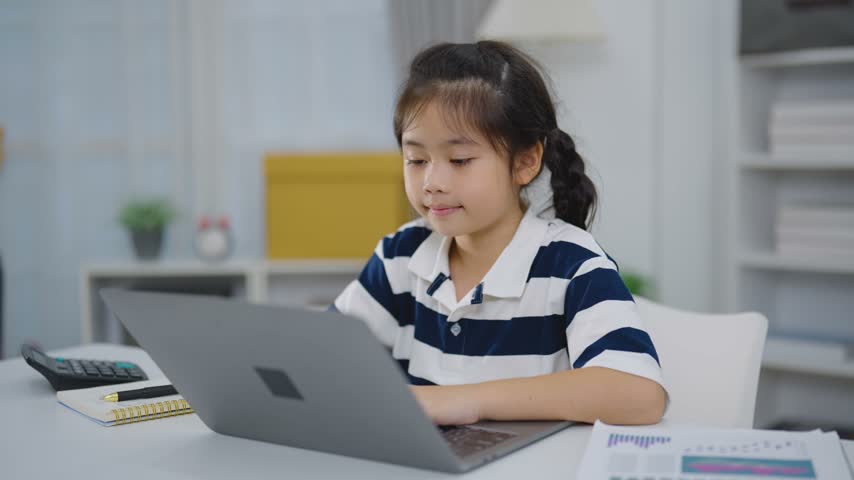 Young girl using laptop computer indoors at home with notebook calculator and chart paper nearby focused on screen