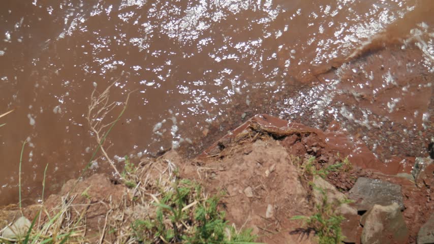Mineral water waves of Lake Shira wash against the rugged red sandstone cliff shoreline with green grass on a sunny summer day, filmed in a high-angle static shot.