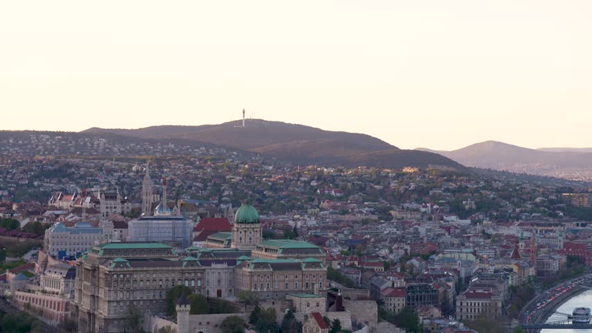 Panoramic View of Buda Castle and Szechenyi Chain Bridge Across Danube River at Sunset in Budapest Cityscape, Hungary Budapest, Hungary - April 7, 2026: High-angle panning shot revealing Buda Castle, hills, and the Danube River spanned by the Szechenyi Chain Bridge at sunset. Warm golden hour light glints across historic architecture, rolling topography, and rippling water where tour boats cruise, framed with a pale gradient sky and deep focal depth.