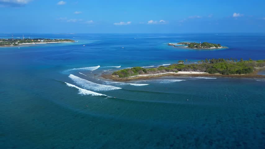 Panoramic drone perspective of tropical coral island surrounded by turquoise ocean waves with surfers in lineup. Ideal visual asset for travel guides and promo.Maldives