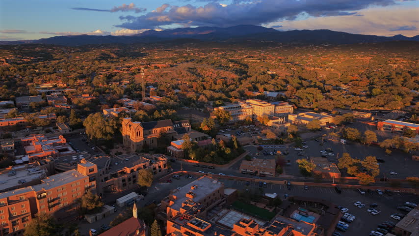 Cathedral Basilica rises over Santa Fe, capital of New Mexico, USA, in low golden sun. Aerial of adobe roofs, parking lots, a hotel pool, autumn trees and dark clouds over Sangre de Cristo Mountains.