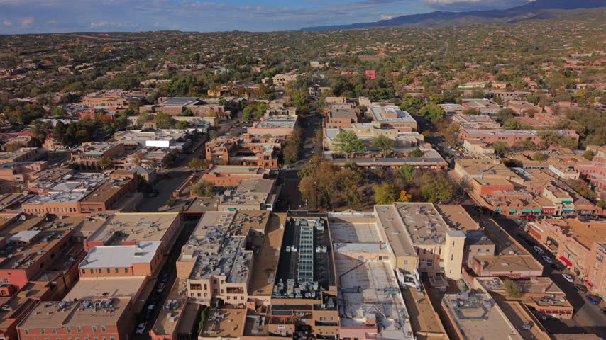 Santa Fe Plaza with autumn trees in the capital of New Mexico, USA. Aerial flyover above flat adobe rooftops and a skylight, Palace of the Governors portal, mountains and cloudy sky in afternoon sun.