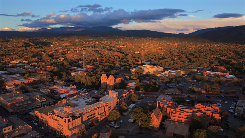 Low evening sun lights downtown Santa Fe, capital of New Mexico, USA. Cathedral Basilica, Loretto Chapel and a terraced adobe hotel above parking lots, dark clouds over Sangre de Cristo Mountains.