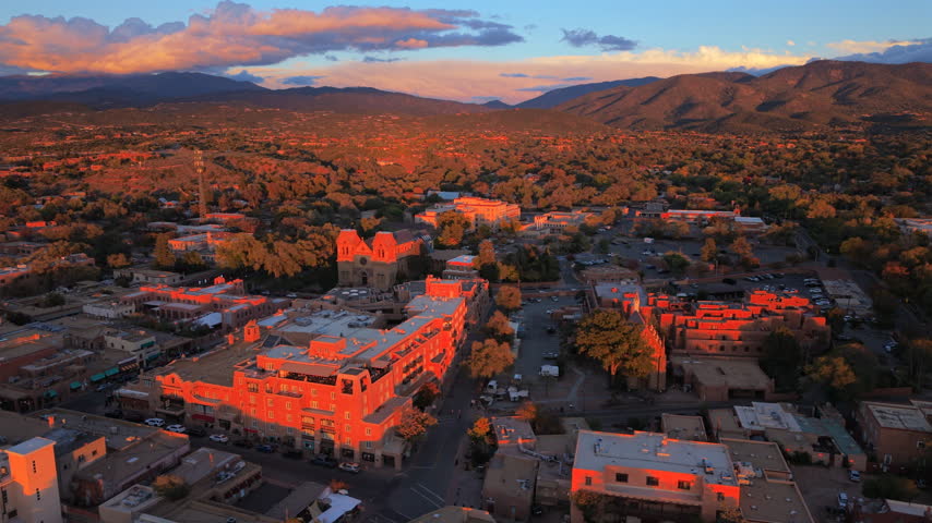 Red sunset glow on terraced adobe hotel and San Loretto chapel Santa Fe, capital of New Mexico, USA. Aerial evening view of Cathedral Basilica, autumn trees and pink clouds over dark mountains.