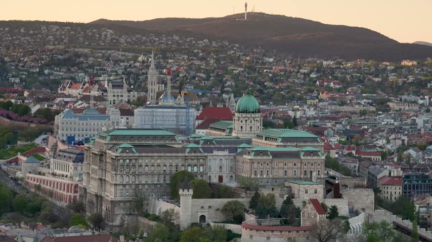 Budapest Cityscape Panning Shot From Buda Castle to Danube River and Szechenyi Chain Bridge at Sunset With Mountain Horizon Budapest, Hungary - April 7, 2026: High-angle panoramic camera pan sweeping across the historical Buda Castle and cityscape to the flowing Danube River and Szechenyi Chain Bridge at dusk. Warm golden sunset light washes over classical rooftops, stone embankments, and surrounding hills, creating gentle tonal contrast and wide depth of field.