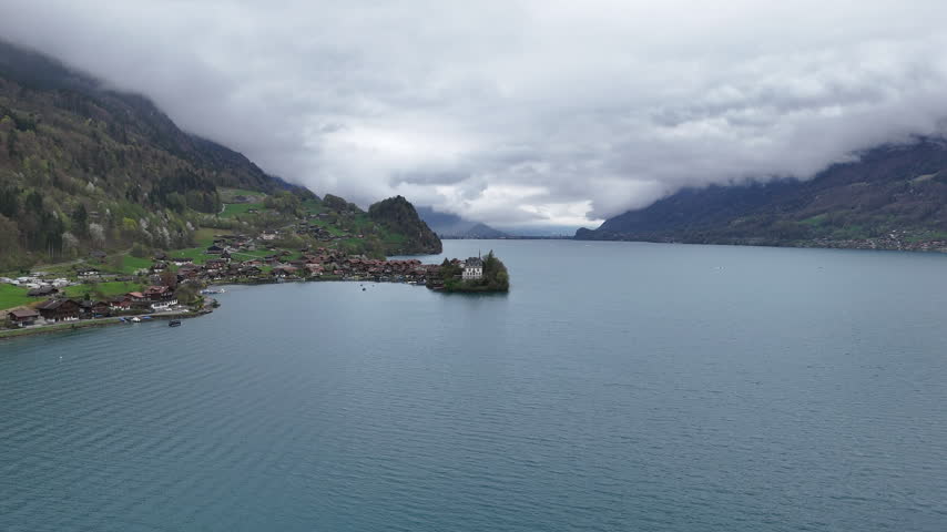 Wide aerial view across a blue alpine lake toward a Swiss village and wooded hills. Low clouds cover the surrounding mountain slopes above the lakeside houses.