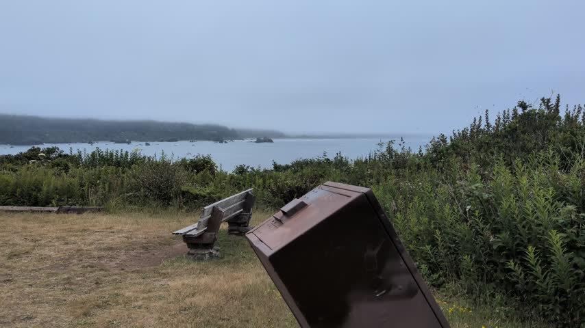 A serene coastal overlook featuring a wooden park bench on a grassy headland, offering sweeping views of a foggy California bay and ocean waters.