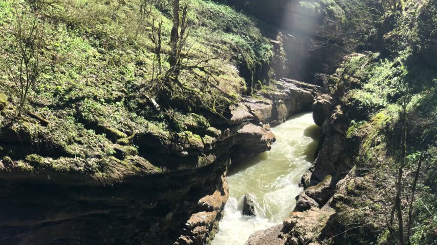 A high-angle view looking down into a deep, narrow gorge where a fast-flowing river rushes through. The water is pale and foamy, cascading over rocky steps in the center of the frame. The gorge is flanked by steep, rugged cliffs covered in lush green vegetation and moss.
