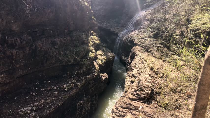 A high-angle view looking down into a narrow, rocky gorge where a stream flows over a small cascade. The water is a pale, milky green, tumbling over a rocky ledge into the channel below. The scene is framed by steep, rugged rock walls on both sides, with patches of green vegetation and moss clinging to the stone. The sunlight creates strong contrasts between the shadowed crevices and the illuminated rock surfaces.