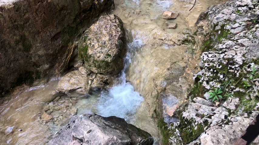 A view looking down into a rocky gorge or crevice where a small, milky-blue stream flows over a rocky ledge, creating a frothy cascade. The scene is framed by rugged, textured rock walls on both sides, with some green vegetation growing in the crevices.