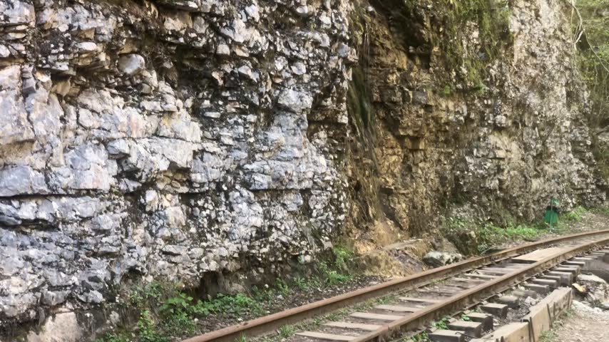 A view along a narrow-gauge railway track running closely alongside a steep, textured rock face. The cliff is composed of jagged, layered stone in shades of grey and brown, with small patches of green vegetation growing from the crevices. The tracks, laid on weathered wooden sleepers, curve gently into the distance on the right side.