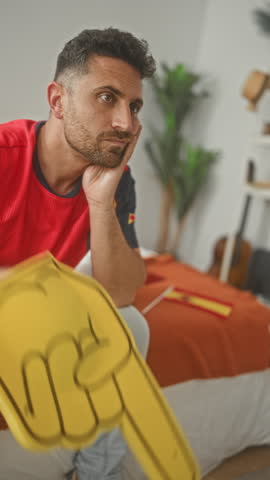 Man resting chin on hand holding yellow foam finger watching soccer match on bed in building; disappointment support.
