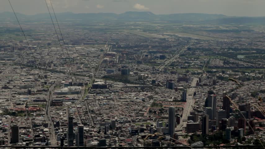 Dense buildings and expanding urban development in Bogotá, Colombia, seen from the hills across the Andean plateau. Electrical cables cross the foreground as atmospheric pollution haze softens the distant city skyline and the hills on the other side of the valley.