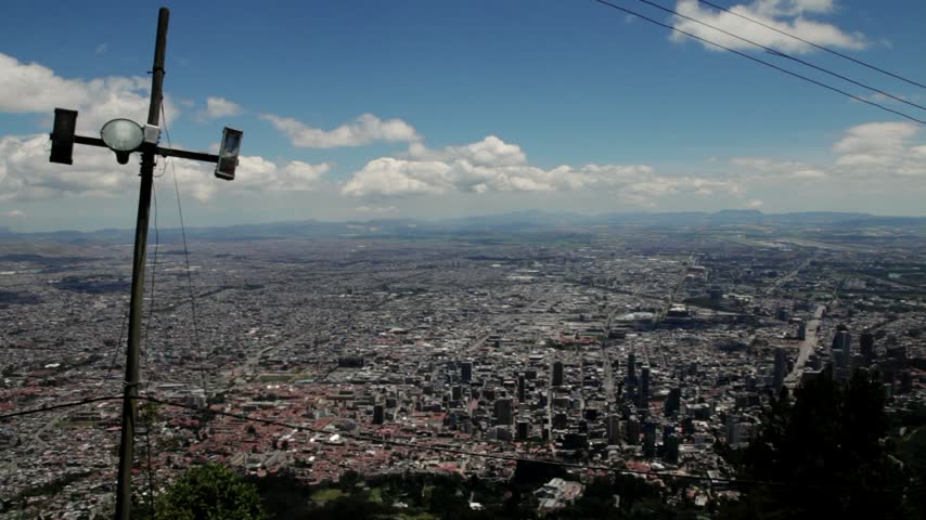Panoramic view of Bogotá, Colombia, spreading across the vast Andean plateau toward the southwestern horizon. A weathered wooden electricity pole and overhead wires contrast with the immense urban landscape under a partly cloudy sky.