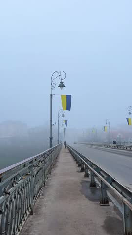 POV walking forward along a city bridge sidewalk in thick morning fog with Ukrainian flags hanging on vintage street lamps and unrecognizable silhouette in the distance.