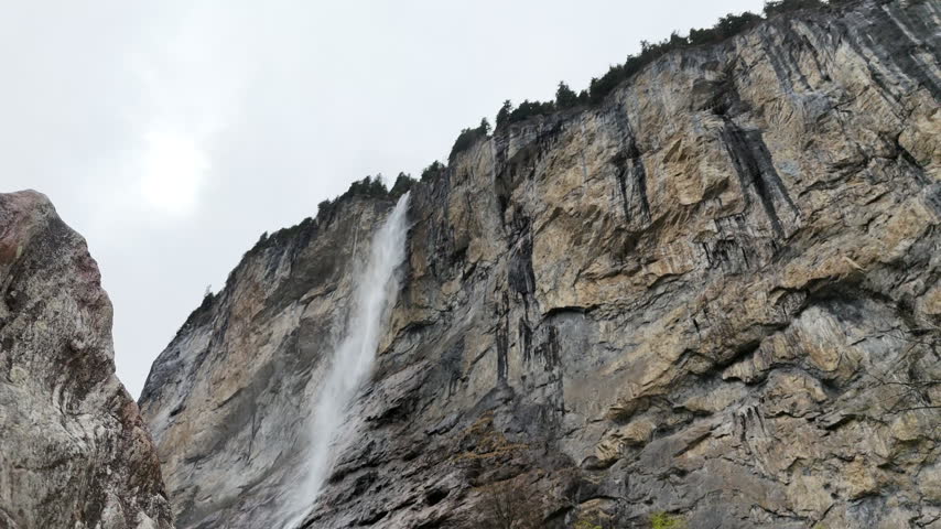 A waterfall descends a tall rock cliff in the Swiss Alps, with a rugged rock outcrop in the foreground and trees lining the cliff edge beneath grey skies.