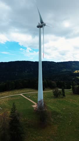 Aerial view of Sainte-Croix wind farm turbine standing in a green meadow with forested hills under a cloudy sky in Sainte-Croix, Vaud, Switzerland.