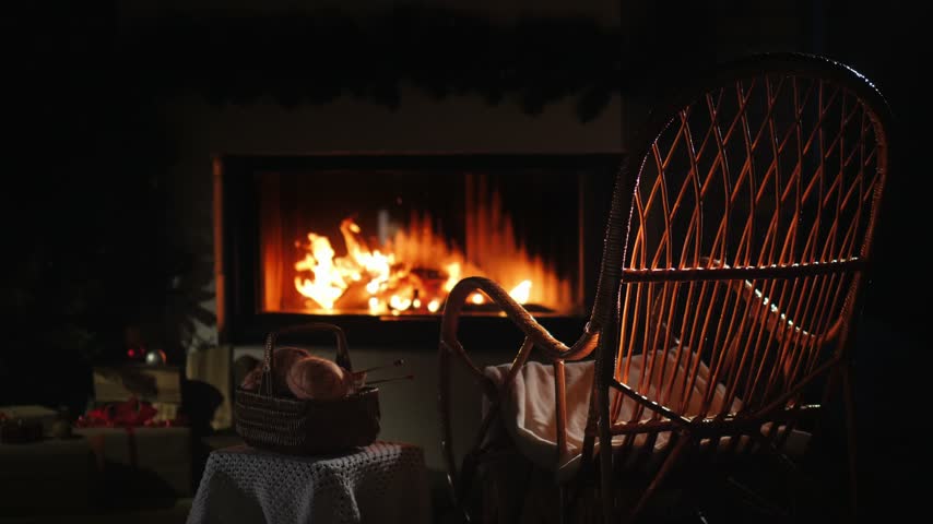Warm living room interior featuring burning fire in hearth empty rocking chair and craft supplies on table at night.
