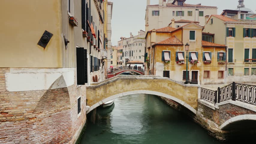 Slow motion camera gliding over Venetian canal with rustic brick architecture and traditional arched bridges at dusk.