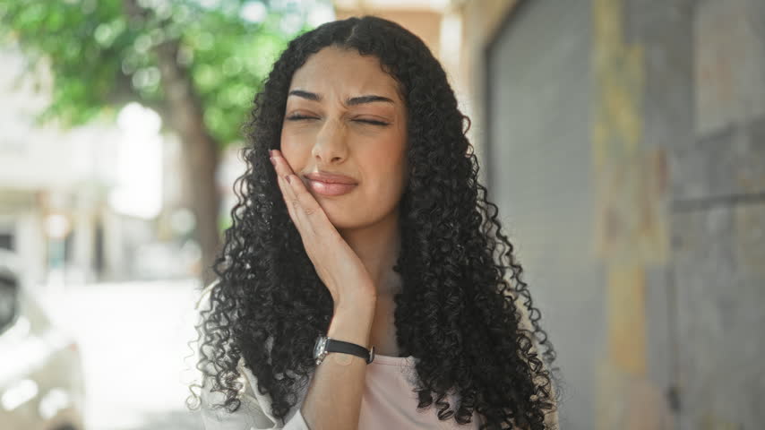 Young arab woman touching cheek with hand in a busy street, wearing wristwatch and light jacket with curly hair framing face; thoughtful doubt.