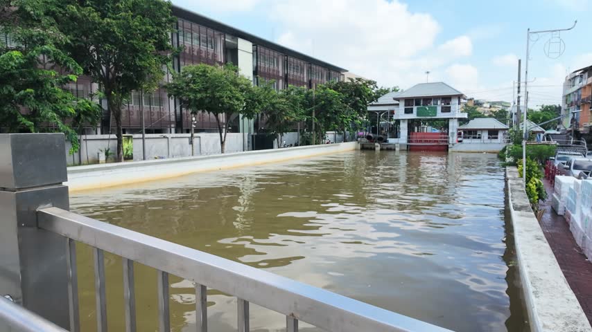 Murky water flows steadily along a concrete urban canal lined with trees and modern buildings.