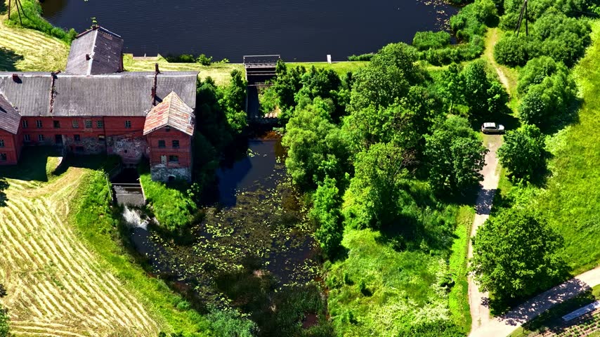 Aerial view of a historic red brick mill building situated beside a stream and pond, surrounded by lush green trees and rural fields in Bene, Dobele Municipality, Latvia.