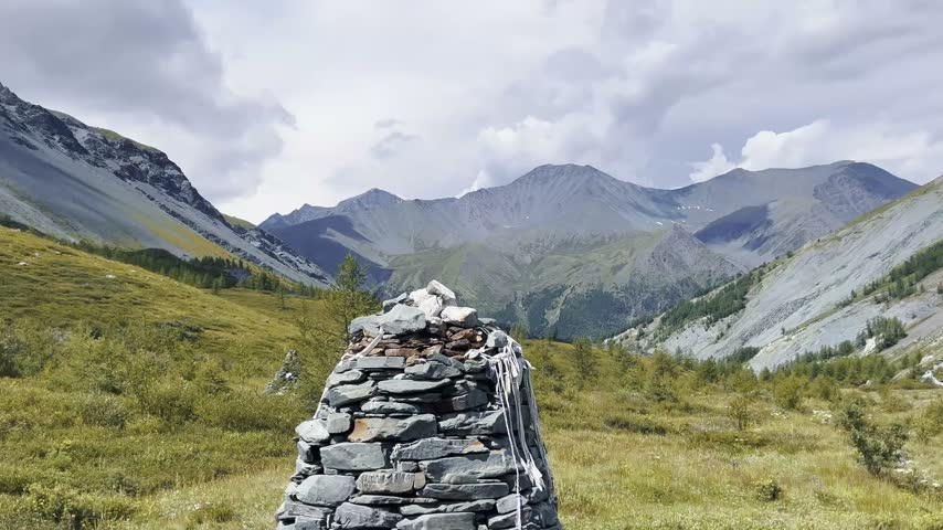 Stone cairn marker standing in alpine meadow, snow capped peaks rising beyond, green slopes descending into misty valleys under diffused cloud cover, establishing remote high altitude terrain.