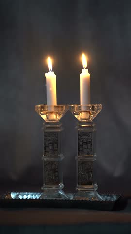 Vertical video of two white Shabbat candles burning in crystal candlesticks engraved with a Jerusalem scene, on a silver tray against a dark background.